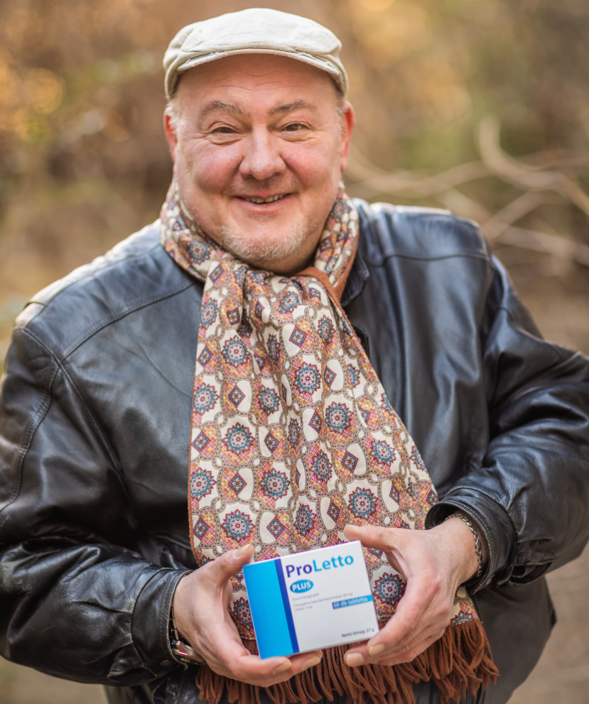 Natural Pharmaceuticals ProLetto® Plus Idős férfi bézs sapkában, sálban és bőrkabátban a ProLetto® Plus gyógyszer dobozát tartja egy fa mellett kültéri környezetben.