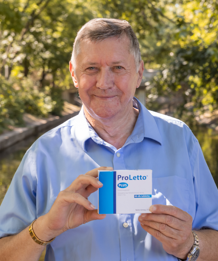 Natural Pharmaceuticals ProLetto® Plus Mosolygó idős férfi áll szabadban, fejhallgatót igazít két kézzel, zöld park háttérrel.